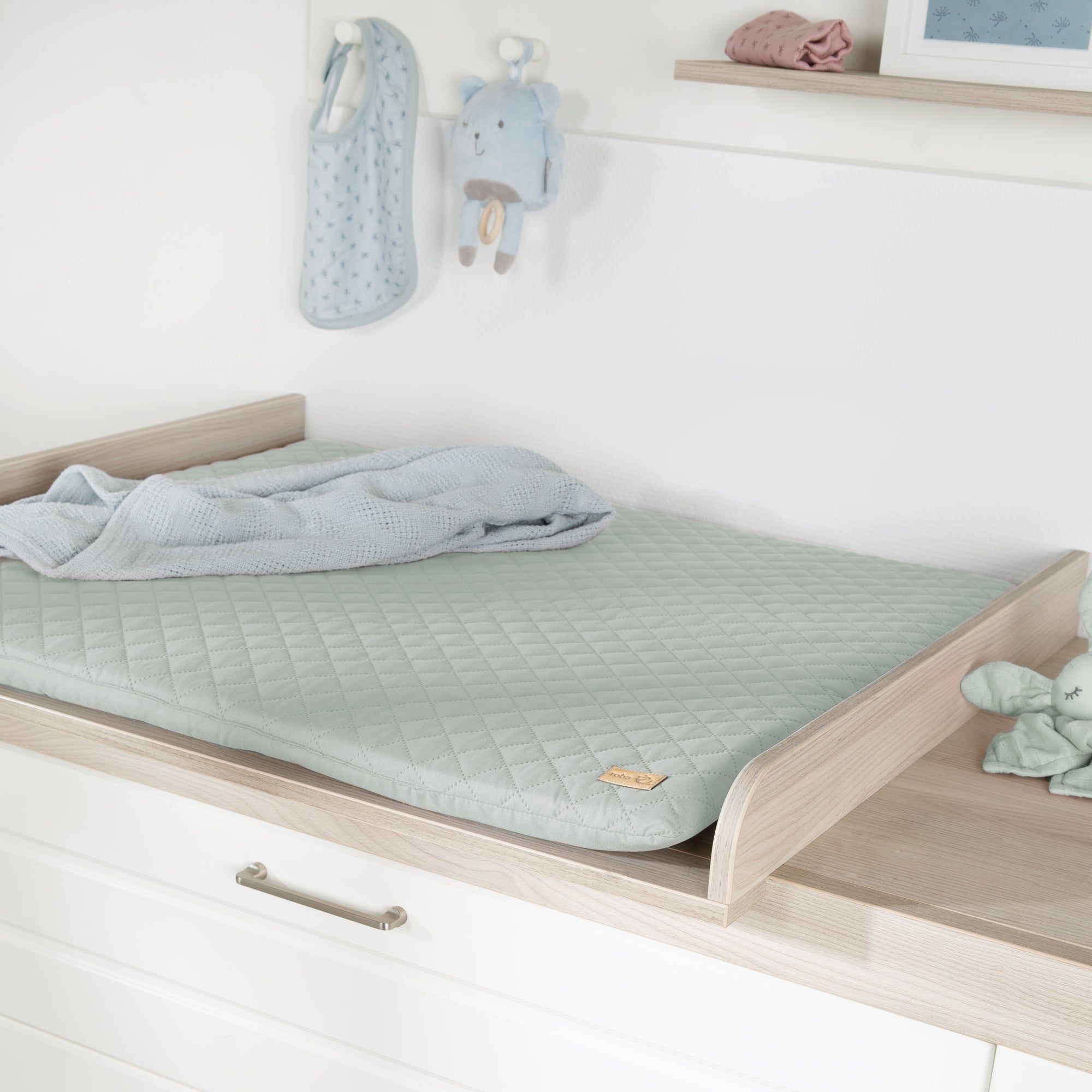Wickelauflage auf Wickeltisch. Hellgrüne, gesteppte Auflage. Holzrahmen. Baby-Accessoires im Hintergrund.