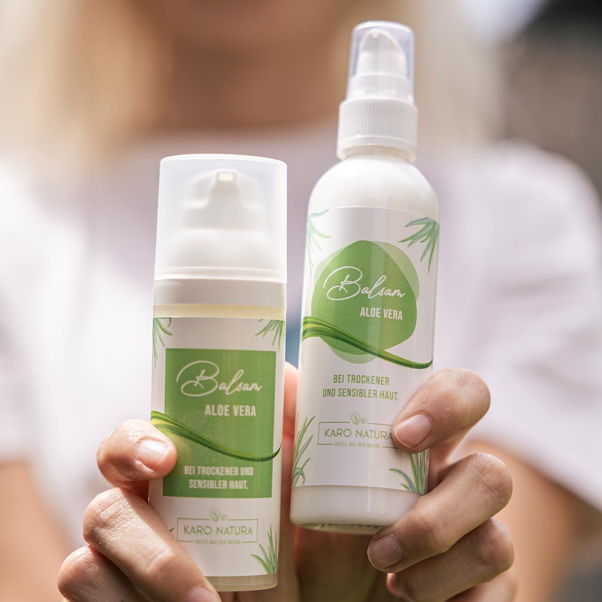 Zwei weiße Balsamflaschen mit "Balsam Aloe Vera" und "Karo Natura" Etiketten. Eine Flasche mit Sprühkopf, eine mit Pumpkopf. Text für trockene und sensible Haut.