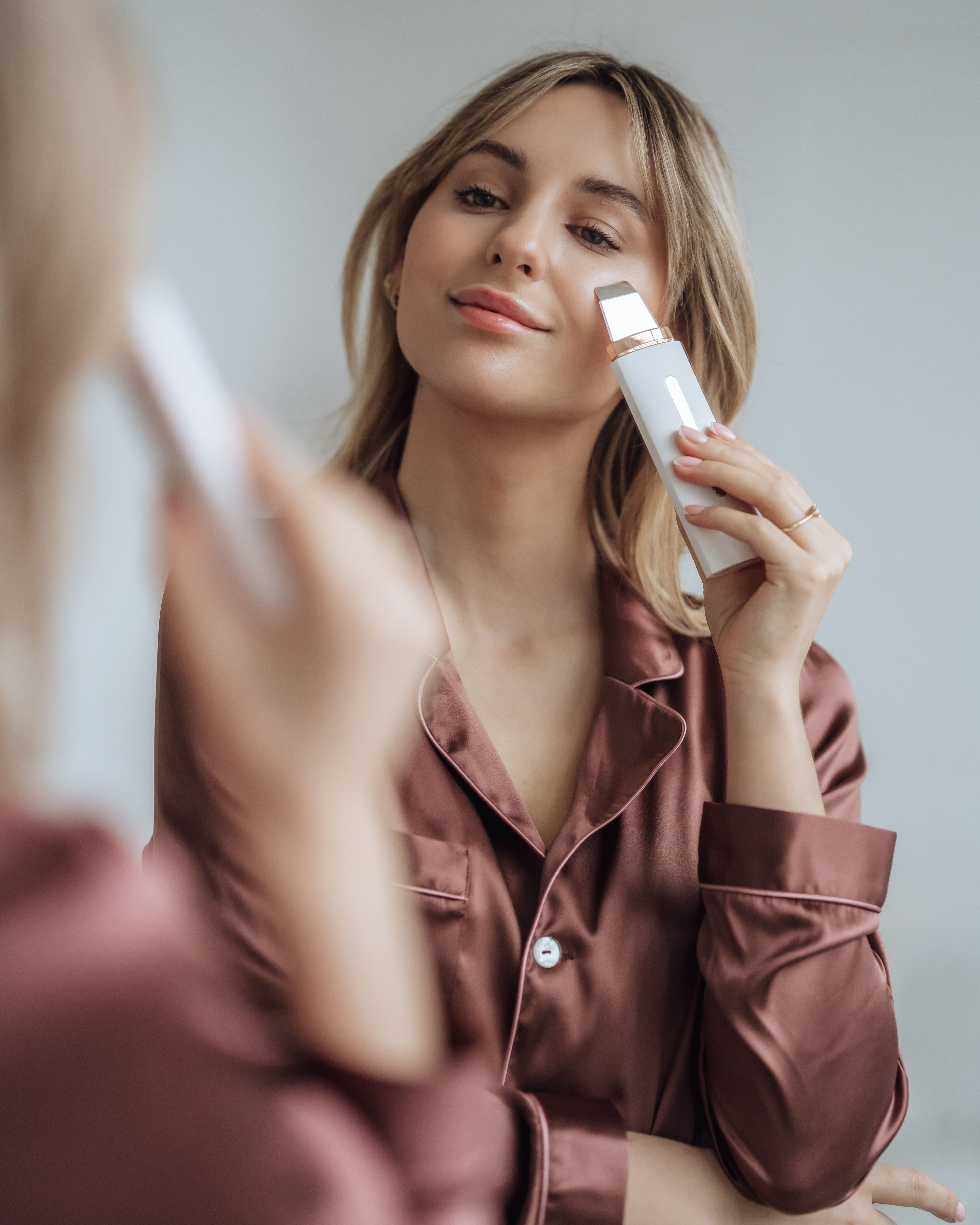 Frau im Spiegel, hält ein weißes BEAUTIFLY B-Scrub Pro Slim Ultraschall Peeling. Sie berührt ihr Gesicht.