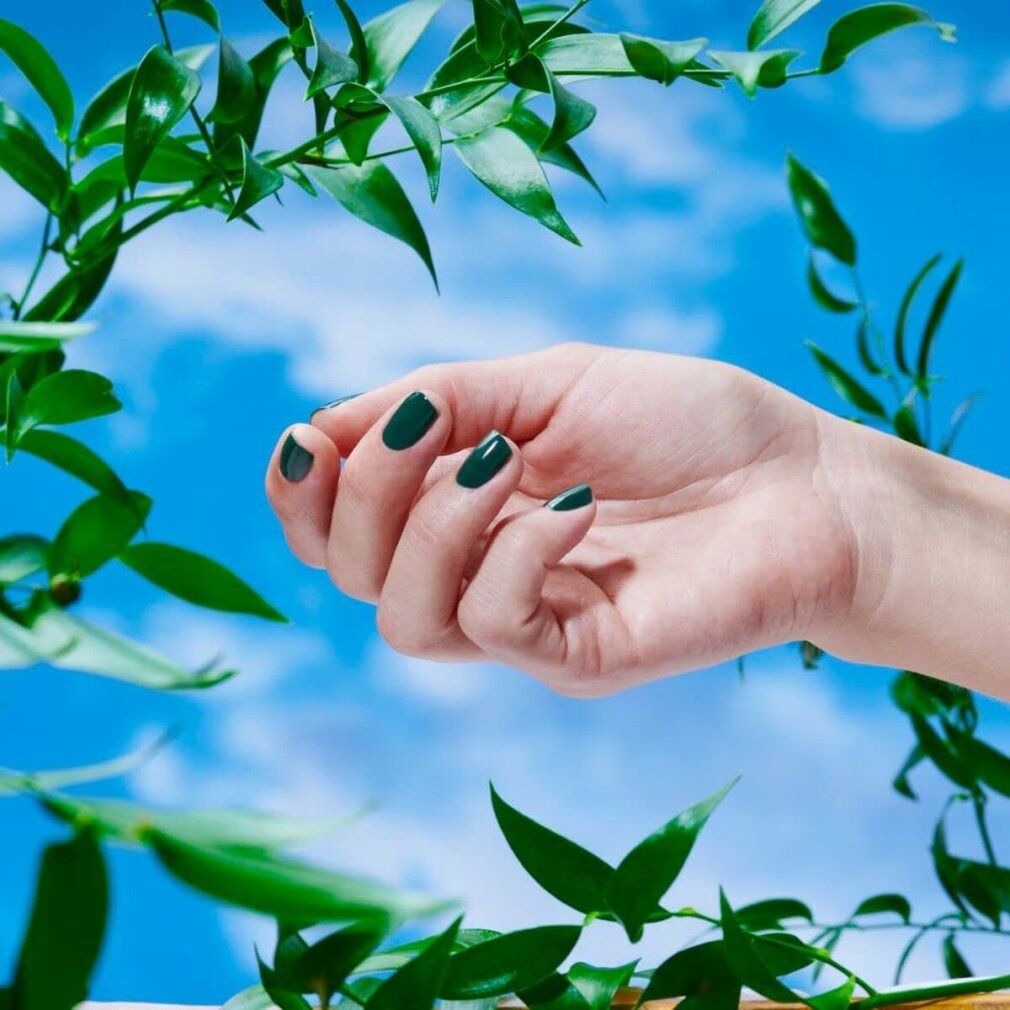 Hand mit lackierten, oval geformten Nägeln in Grün. Blauer Himmel im Hintergrund.
