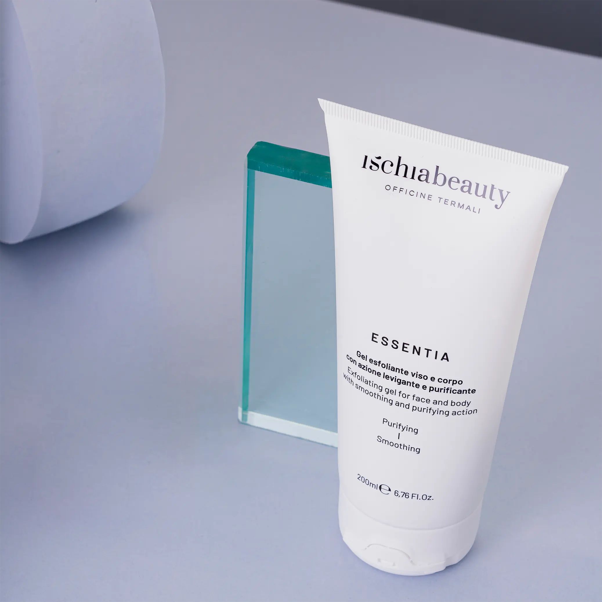 Weiße Tube mit Produktbeschriftung. Text: Ischiabeauty, Essentia, Gel esfoliante viso e corpo. Auf blauem Hintergrund, neben Glasobjekt.