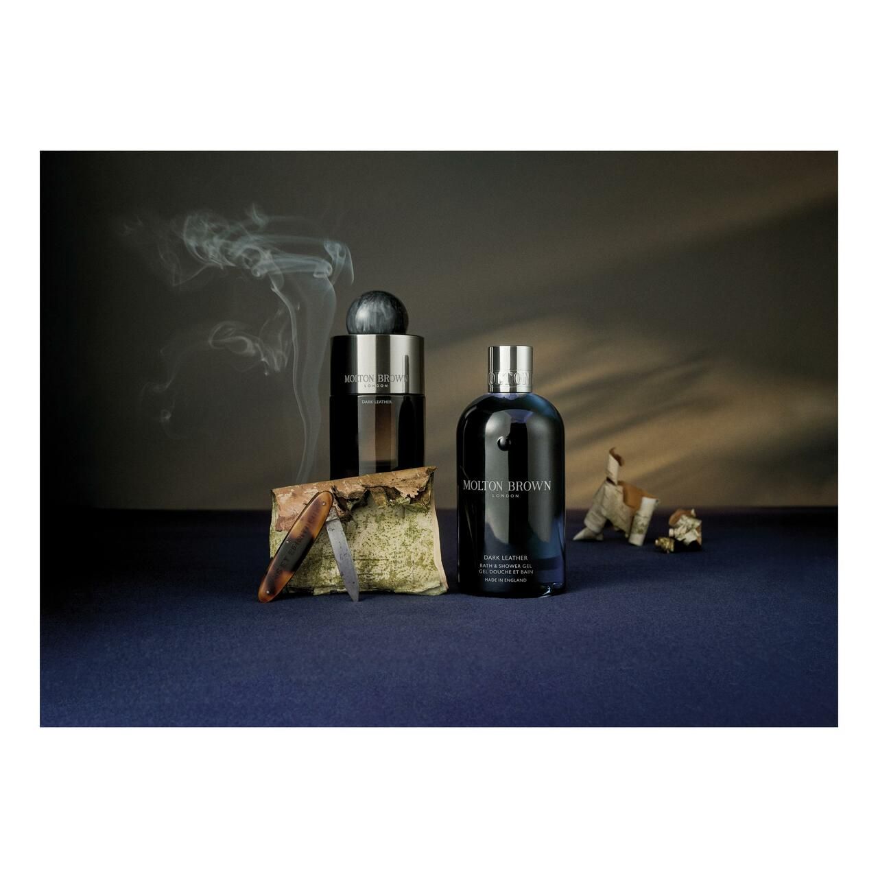 Dunkelblaue Flasche, neben einer weiteren Flasche und Accessoires auf blauem Untergrund. Rauch im Hintergrund.