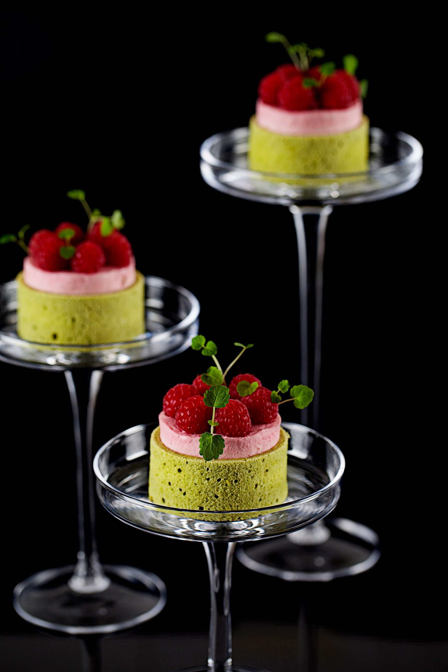Drei kleine Kuchen auf Glasfüßen mit Himbeeren und Minze auf schwarzem Untergrund.