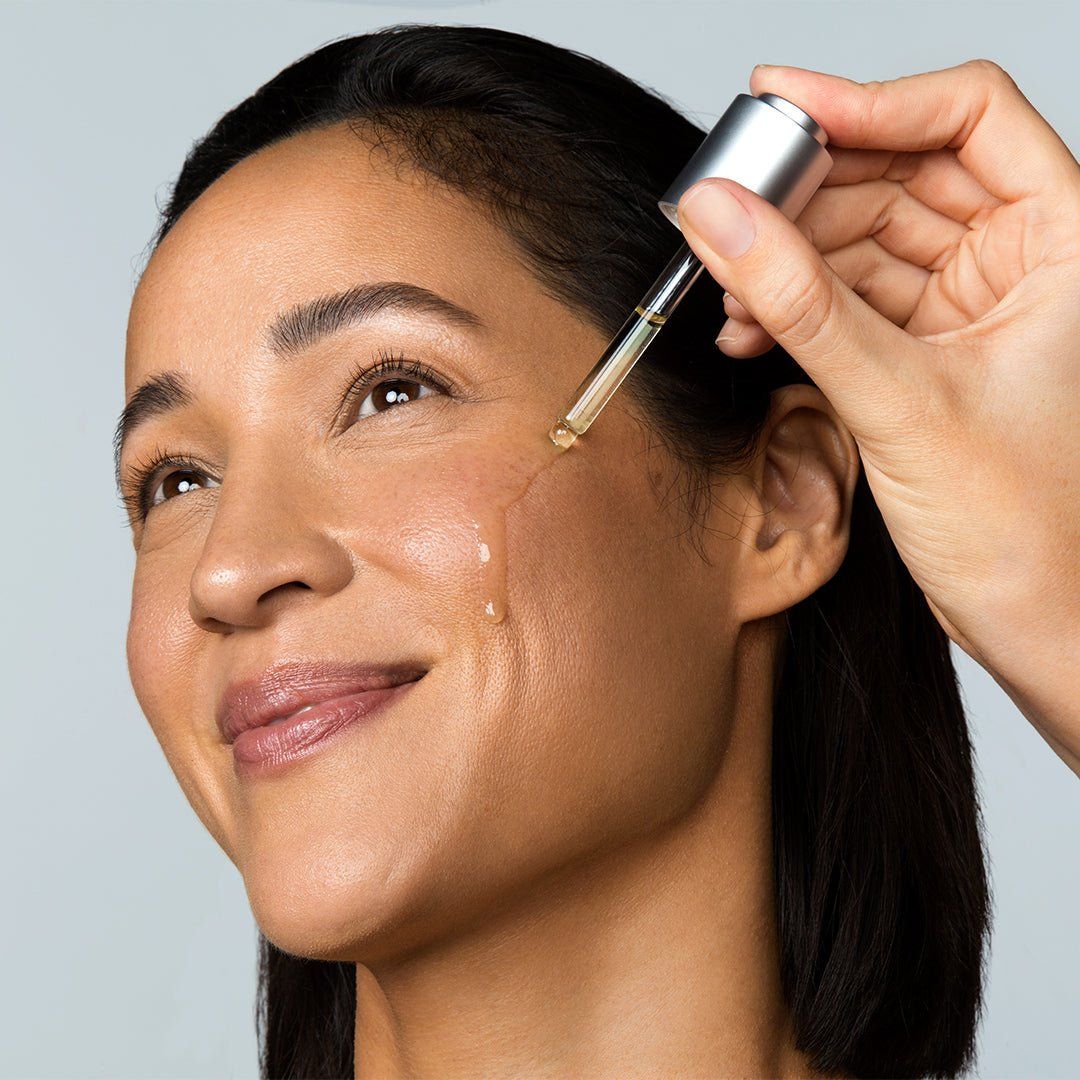 Gesicht einer Frau, auf die ein Produkt aufgetragen wird. Eine Hand hält eine Pipette mit Flüssigkeit, die auf die Haut tropft.