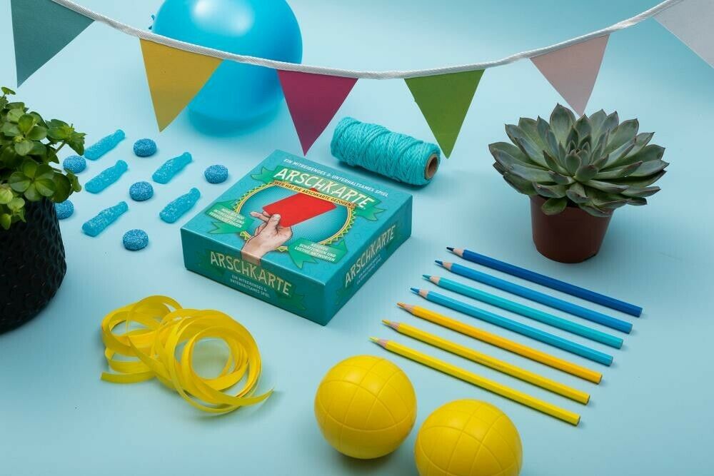 Spielbox "Arschkarte" mit roter Karte, umgeben von Dekoration: Girlande, Ballons, Pflanzen, Stiften.