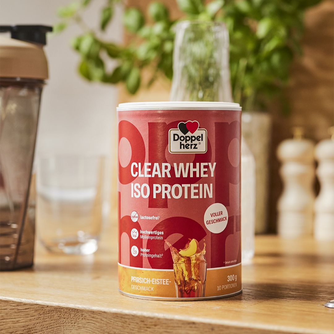 Dose Doppelherz CLEAR WHEY ISO PROTEIN mit Pfirsich-Eistee Geschmack. Steht auf einer Holzoberfläche neben einem Shaker und einem Glas Wasser.