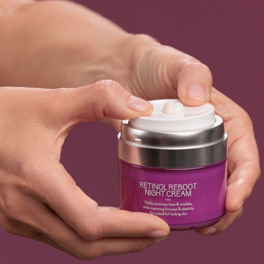 Hände halten ein violettes Cremeglas mit silbernem Deckel. Aufschrift: RETINOL REBOOT NIGHT CREAM. Text: Minimiert Linien & Falten, verbessert die Hautelastizität.
