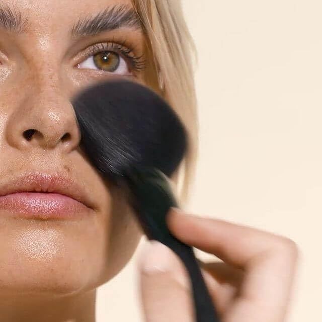 Person mit Pinsel, der Puder auf das Gesicht aufträgt. Nahaufnahme. Heller Hintergrund.