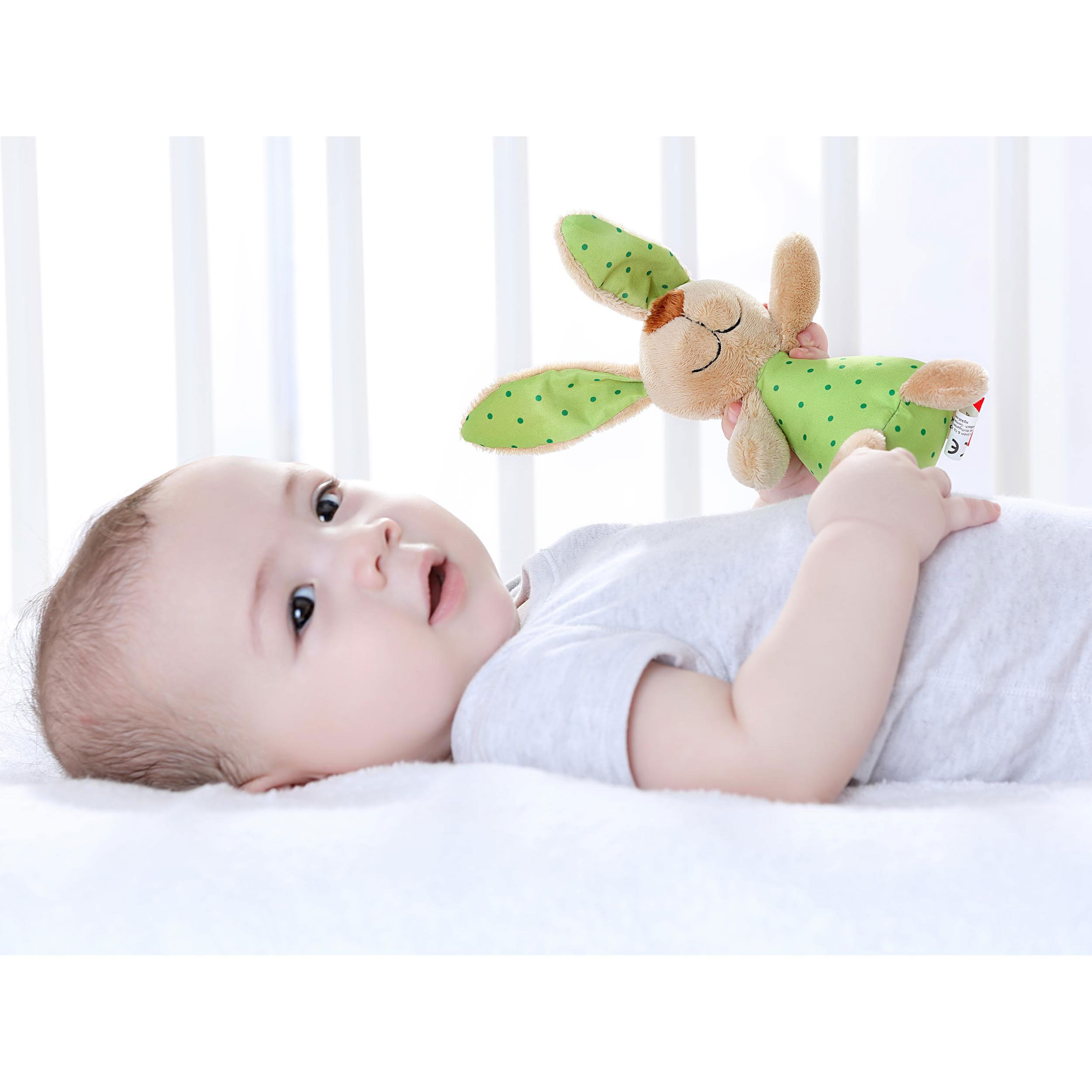 Baby liegt auf dem Rücken und hält einen Stofftier-Hasen mit grünen Punkten.
