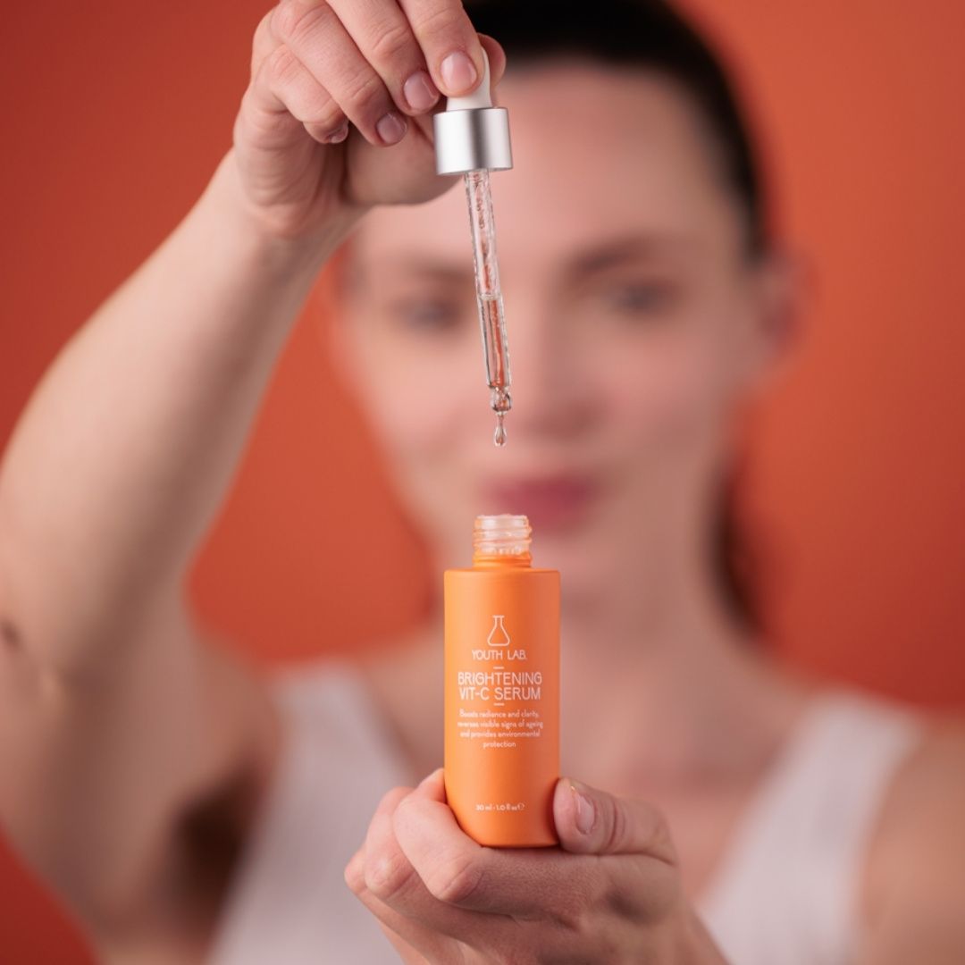 Frau hält Flasche und Pipette. Flasche orange, Aufschrift: YOUTH LAB Brightening Vit-C Serum. Hintergrund orange.