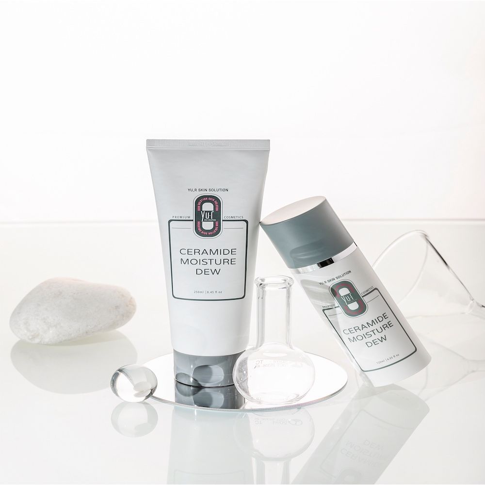 Zwei Produkte: Tube und Flasche. Aufschrift: Ceramide Moisture Dew. Marke: Yu.R Skin Solution. Auf Glasplatte mit Glaswaren und Kieselsteinen.