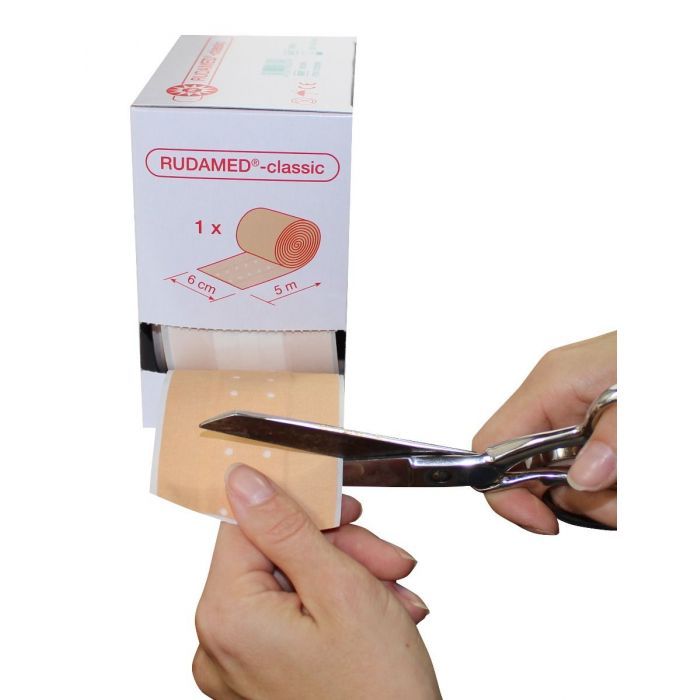 Rudamed-classic Wundpflaster-Verpackung. Schere schneidet Pflaster ab. Aufschrift: Wundschnellverband, 6 cm x 5 m.