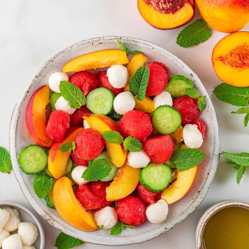 Salat mit Wassermelone, Gurke, Pfirsich, Mozzarella und Minze. In einer Schüssel. Mit Dressing und Mozzarella-Kugeln daneben.