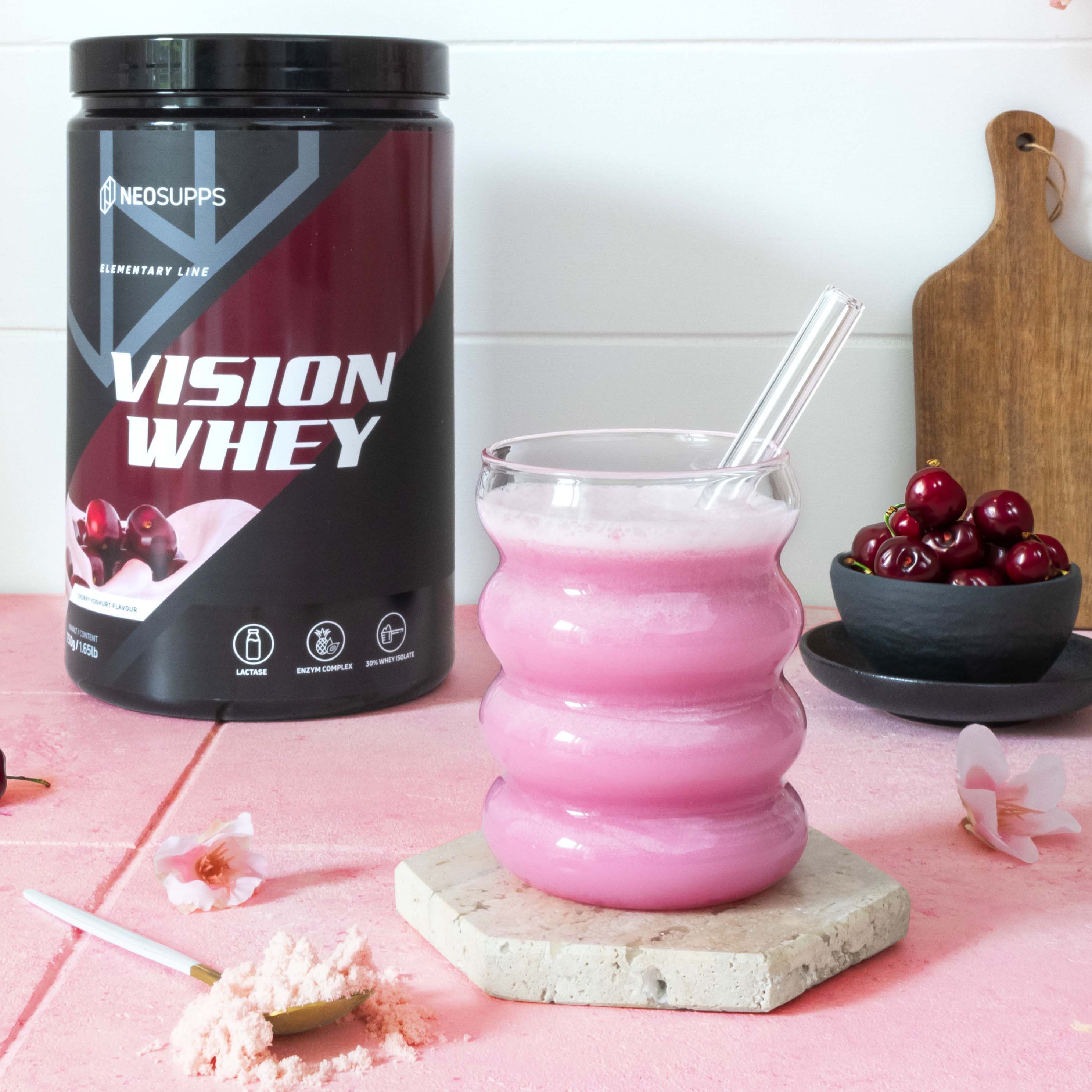 Glas mit rosa Flüssigkeit und Strohhalm. Dose Vision Whey daneben. Schwarze Dose mit rotem Etikett. Kirschen und Löffel daneben.