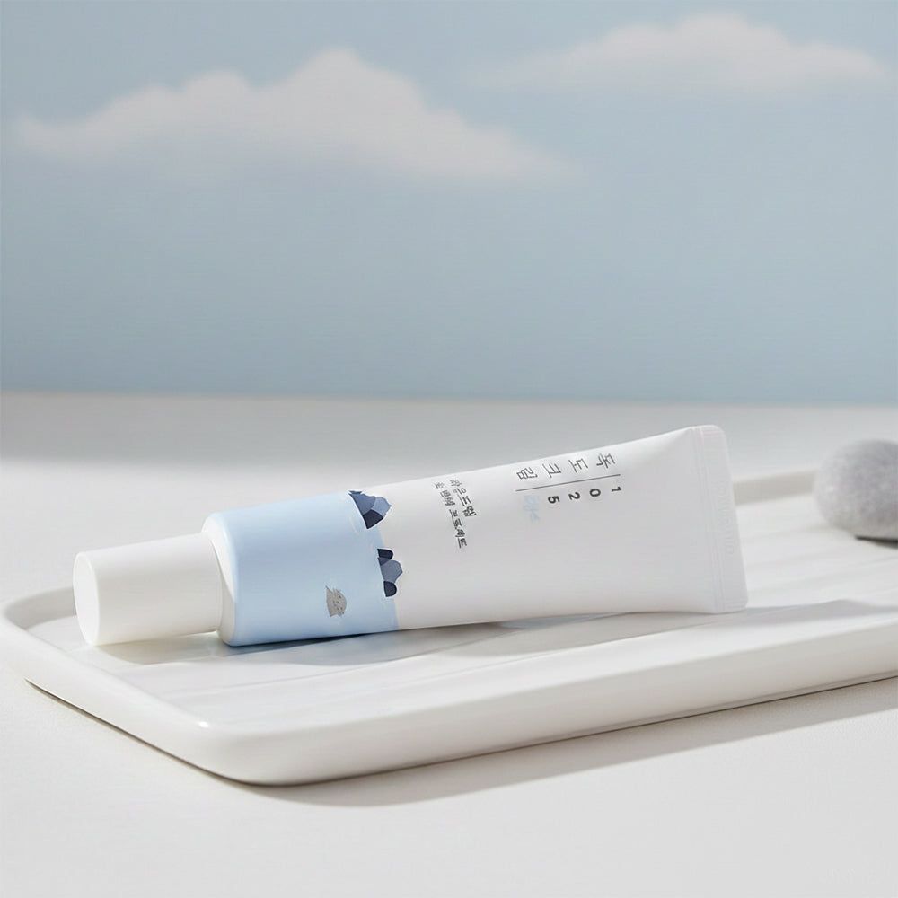 Weiße Tube mit blauer Aufschrift und Illustration. Liegt auf einem weißen Tablett. Hintergrund: Himmel mit Wolken.