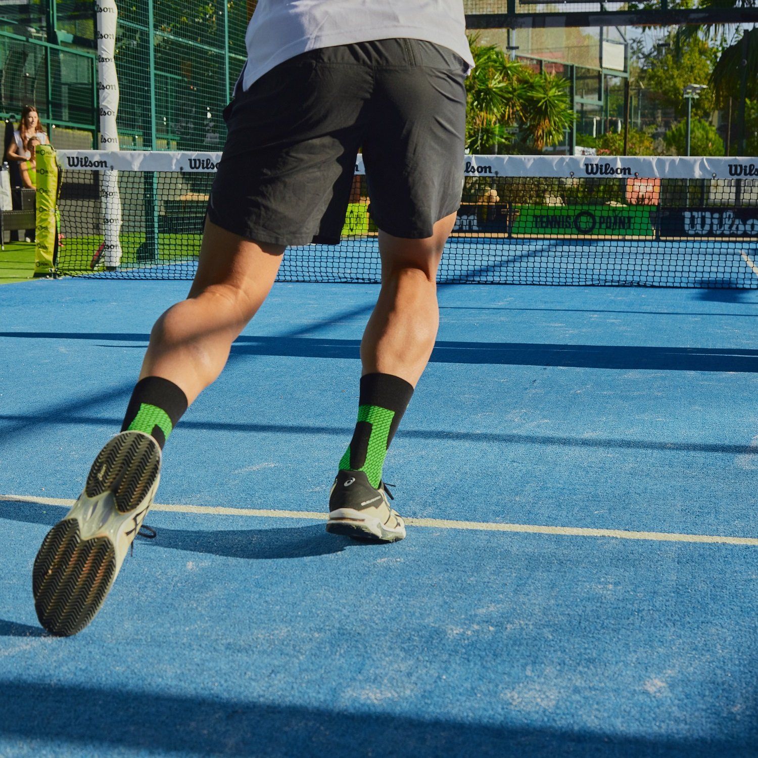 Un uomo gioca a tennis su un campo blu. Indossa pantaloncini neri e calzini stabilizzatori di caviglia Enforma, neri con dettagli verdi. Le sue scarpe da tennis sono in primo piano mentre si muove.