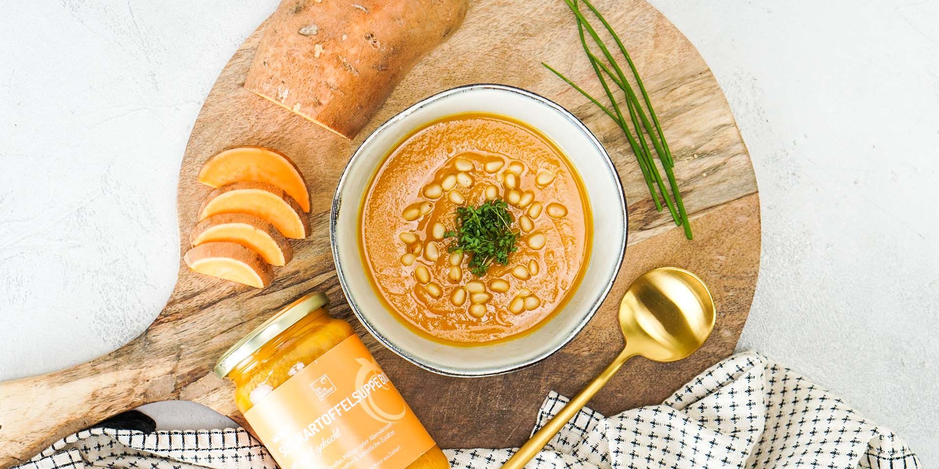 Schüssel mit Süßkartoffelsuppe, Pinienkernen und Kräutern. Daneben Glas Wacker Süßkartoffelsuppe Bio, Holzbrett, Löffel.