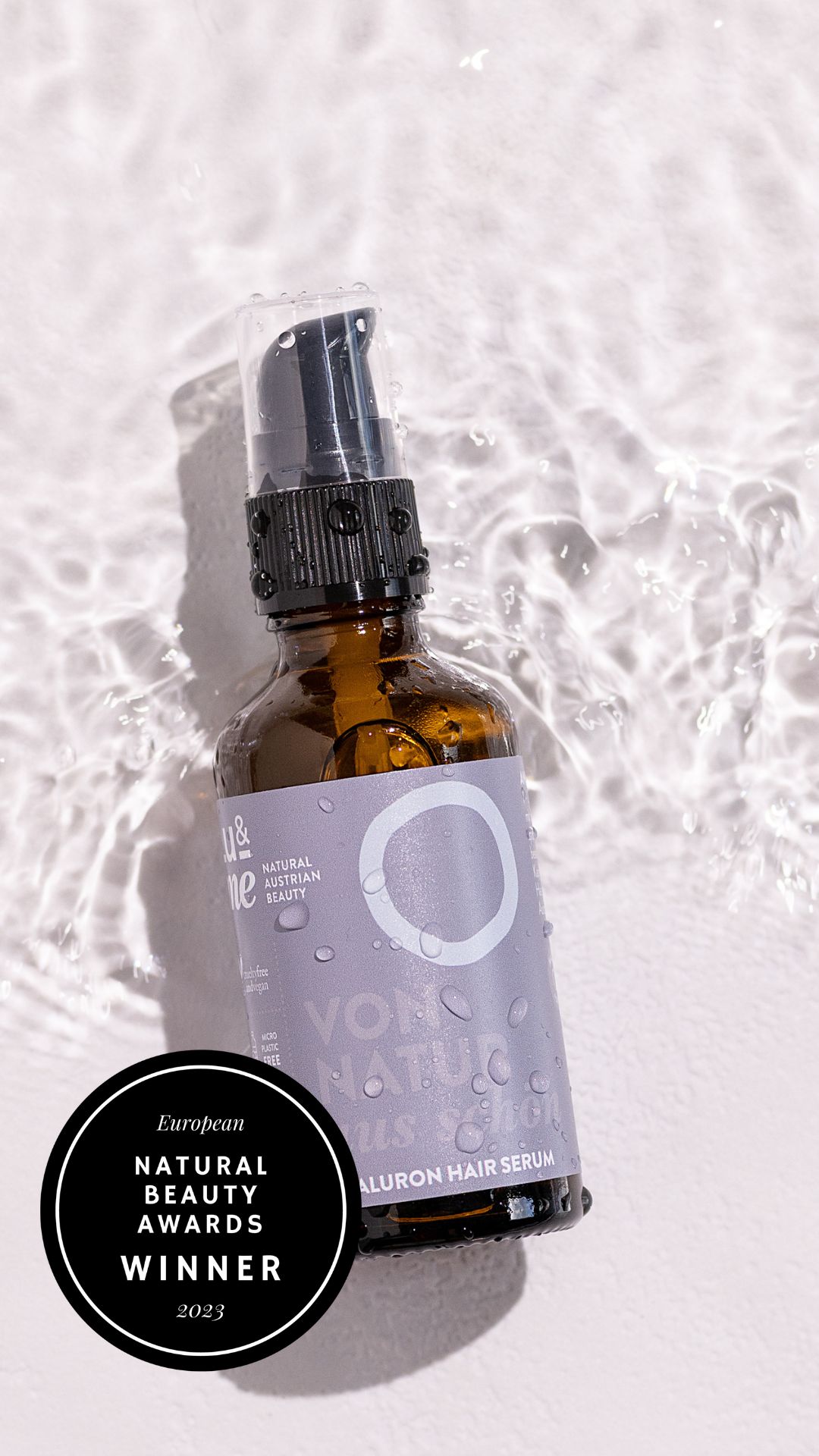 Braune Glasflasche mit schwarzem Sprühkopf, teilweise im Wasser. Etikett mit Logo und Text. Auszeichnung 'European Natural Beauty Awards'.