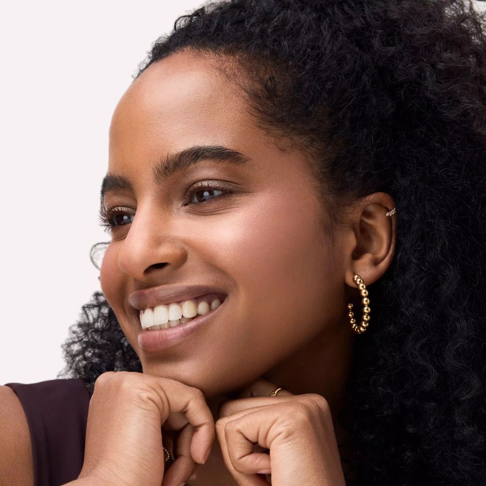 Porträt einer lächelnden Frau mit dunklem Haar und goldenen Ohrringen. Sie stützt ihr Kinn auf ihre Hände.