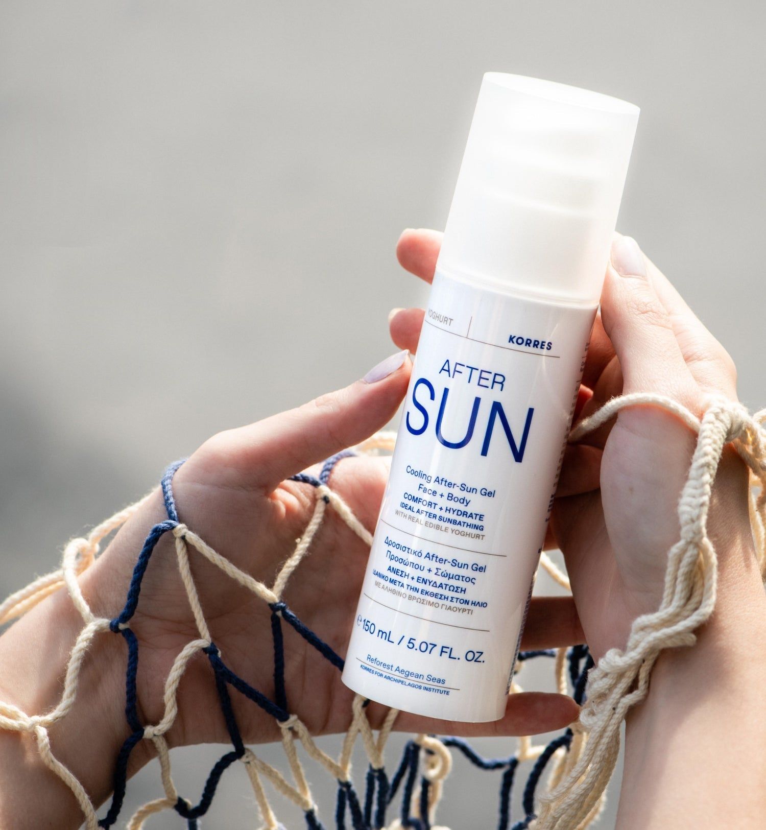Weiße Flasche mit "AFTER SUN"-Aufdruck. Hände halten die Flasche vor einem Netz. Text: "KORRES", "Cooling After-Sun Gel".