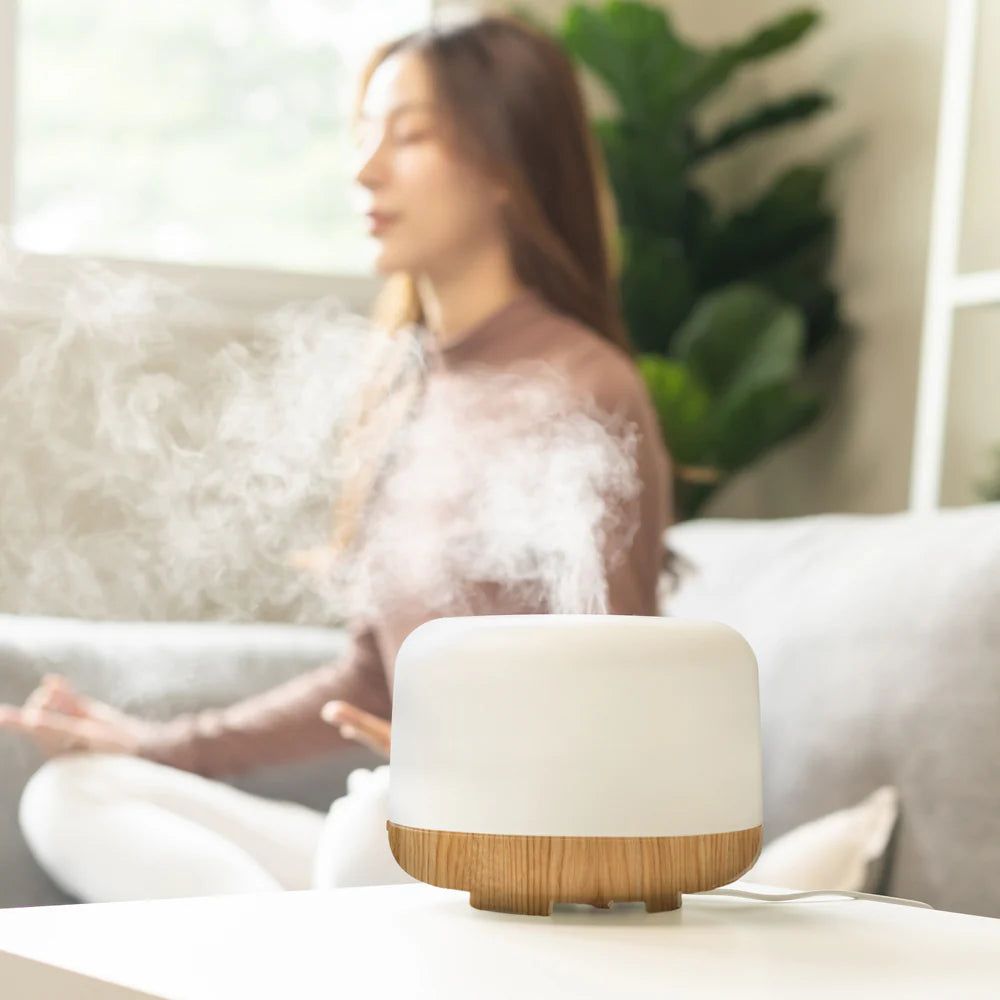 Aroma Diffusor mit Holzmaserung-Basis. Weißer, runder Körper mit Holzbasis. Frau sitzt im Hintergrund. Dampf steigt auf.