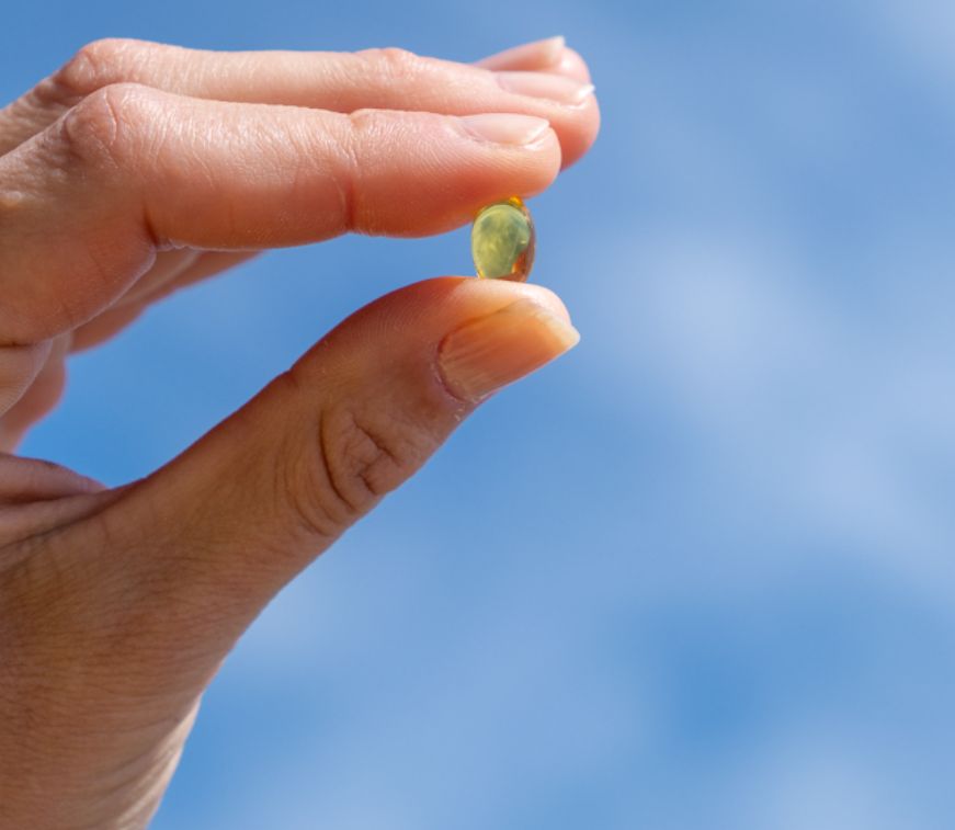 Eine Hand hält ein einzelnes Aavalabs Vitamin D3+K2 Softgel vor blauem Himmel.
