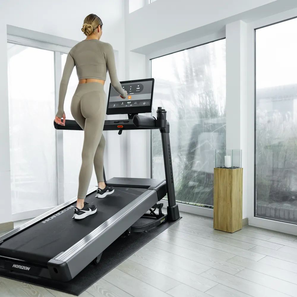 Femme marchant sur un tapis de course. Elle porte des vêtements de sport. Tapis de course noir. Logo Horizon visible. Sur fond blanc.