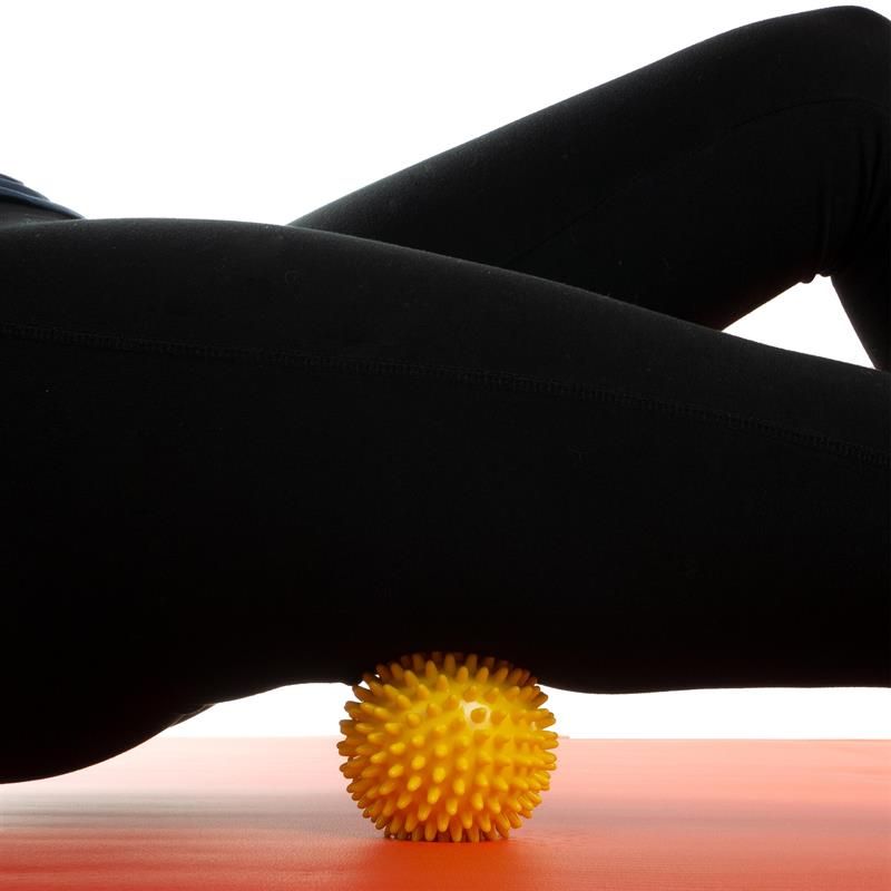 Gelbe Massagekugel unter dem Oberschenkel einer Person, die auf dem Boden liegt. Schwarze Hose.