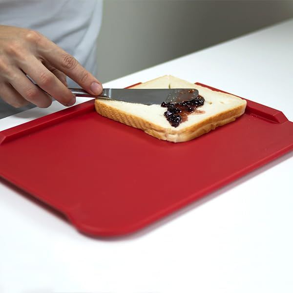 Rotes Essbrettchen. Eine Hand bestreicht Toastbrot mit Marmelade. Das Brot liegt auf dem Brettchen. Rechteckige Form mit abgerundeten Ecken.