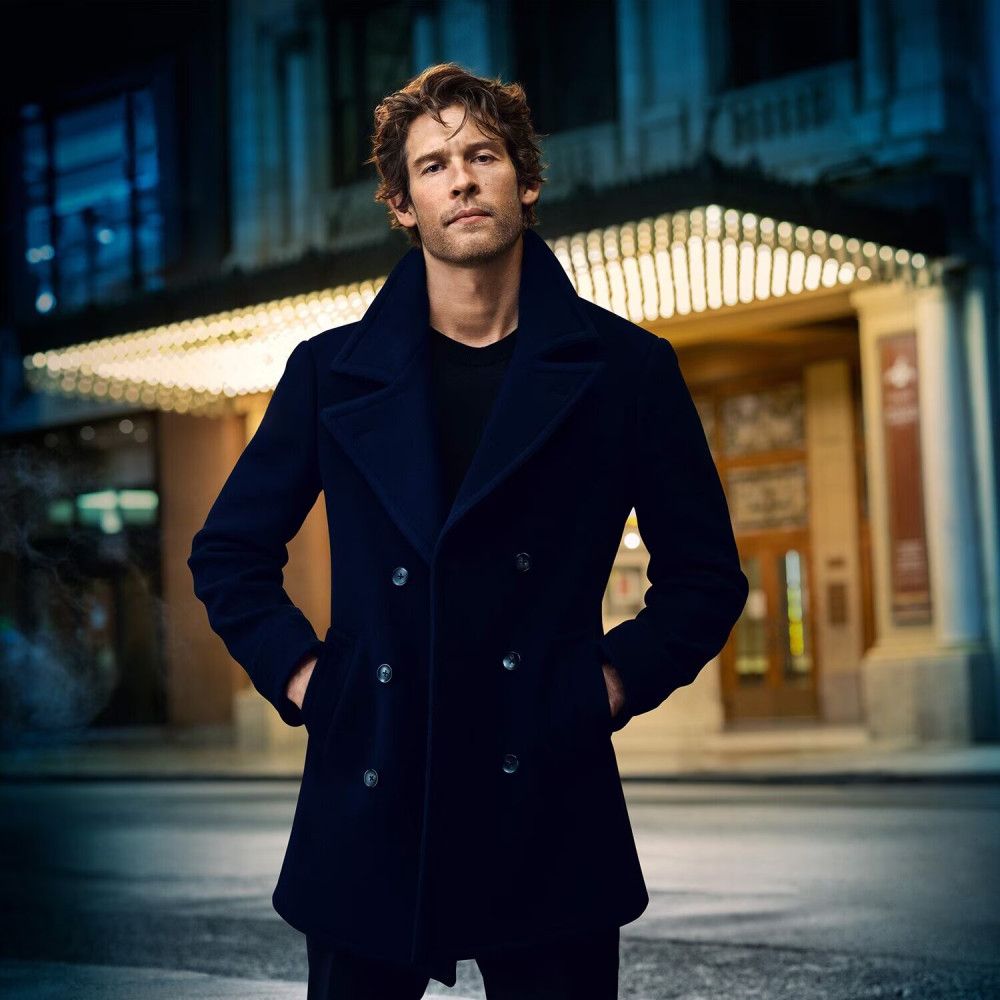 Mann in dunkelblauem Mantel vor einem Gebäude. Hände in den Hosentaschen.