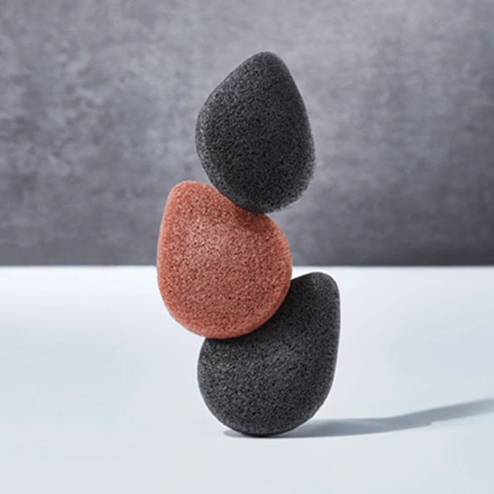 Drei Konjac-Schwämme in Balance. Zwei schwarz, einer rotbraun. Tropfenform.