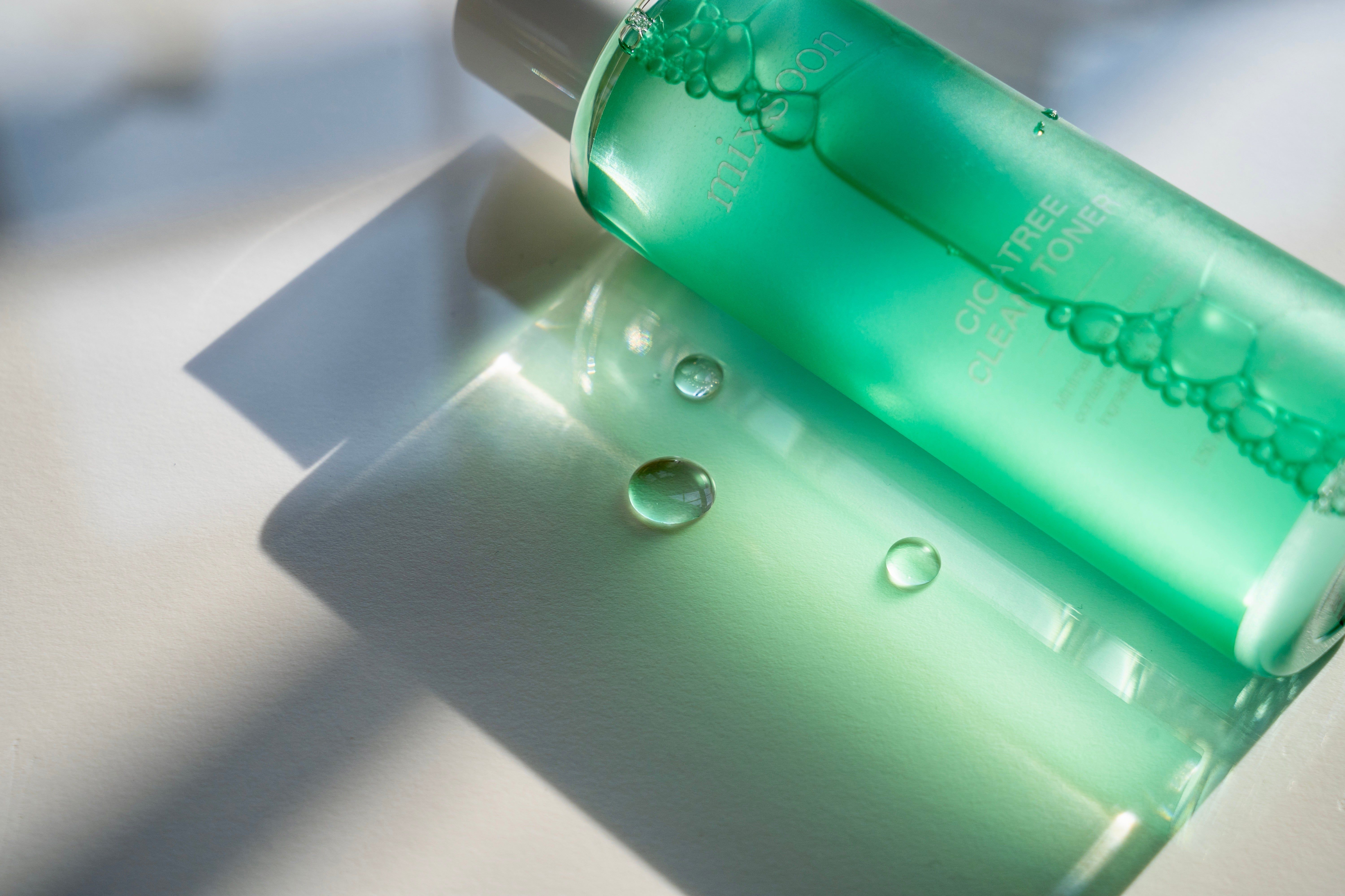Grüne Flasche und Wassertropfen. Aufschrift: mixsoon, Cicatree Clean Toner. Schatten. Hintergrund: hell.