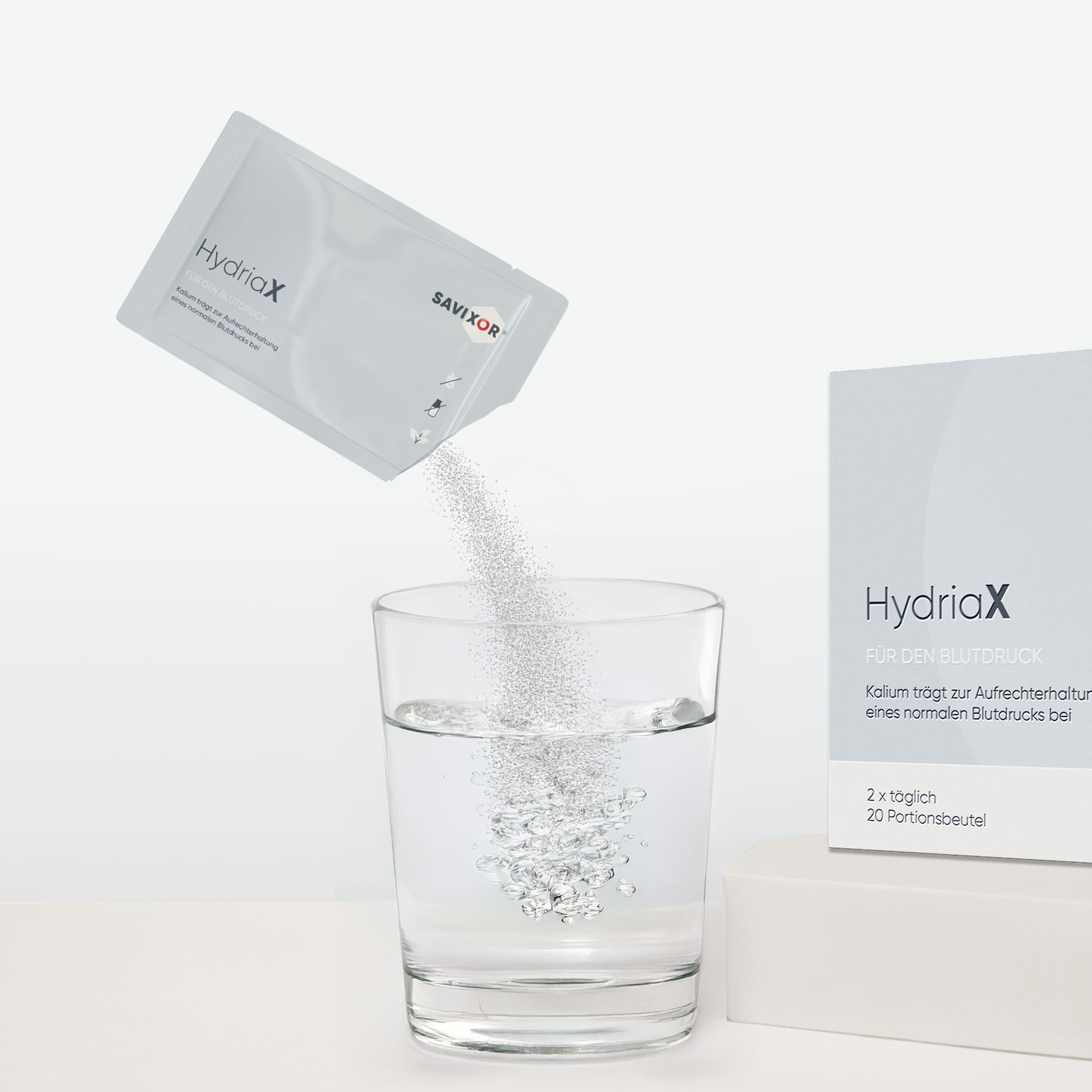 Savixor HydriaX Elektrolyte Pulver, Sachet. Pulver wird in ein Glas Wasser gegossen. Daneben eine Packung und ein Glas Wasser.