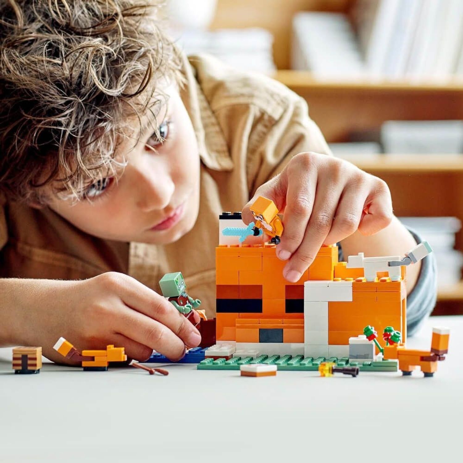 Kind baut LEGO Minecraft 21178. Orangefarbenes Gebäude mit weißen Akzenten. Figuren: Fuchs, Zombie, Füchse. Kind im Vordergrund.