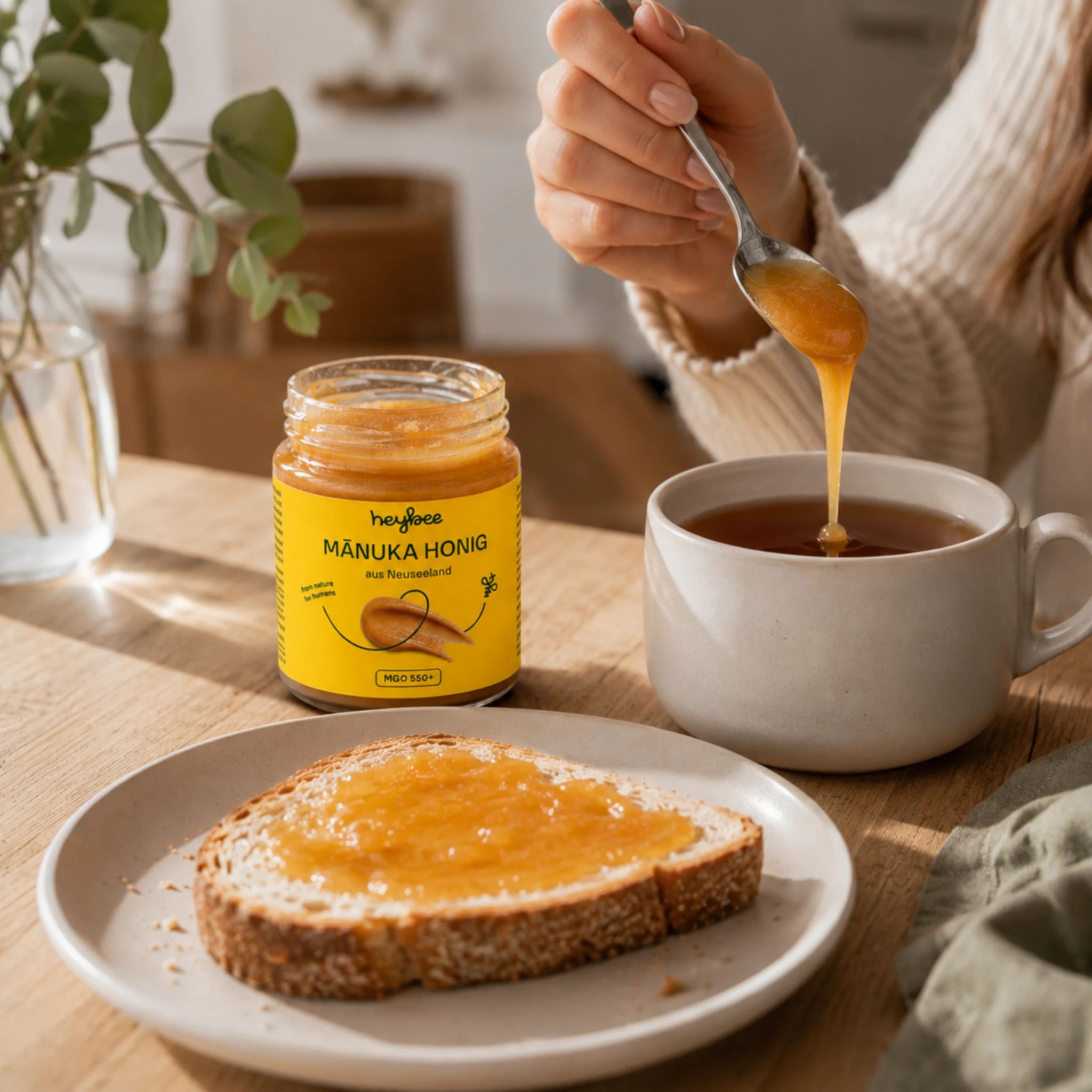 Glas Manuka Honig mit gelbem Etikett, Löffel mit Honig über einer Tasse Tee und Toast.