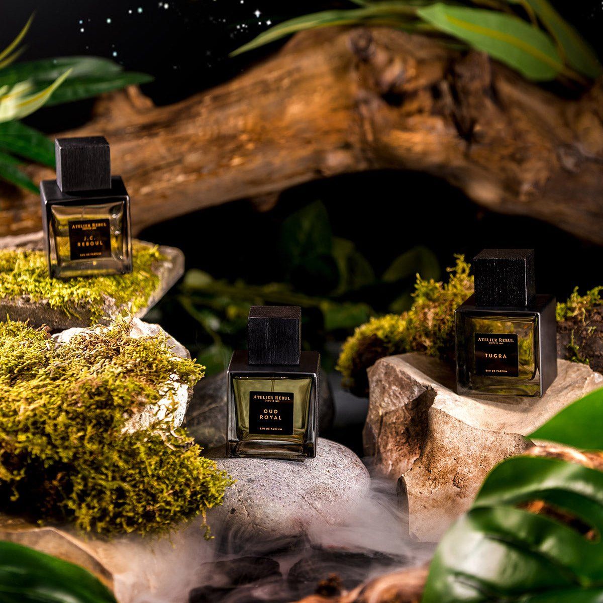 Drei Parfümflakons mit schwarzen Deckeln auf Steinen und Moos. Aufschrift: Oud Royal, Le Désir, Tigra. Atelier Rebul Logo.