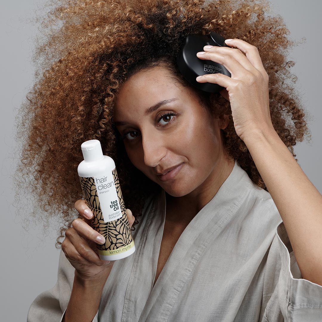 Frau mit Kopfmassagebürste und Flasche. Frau trägt Bademantel. Flasche: Hair Clean Tea Tree Oil.