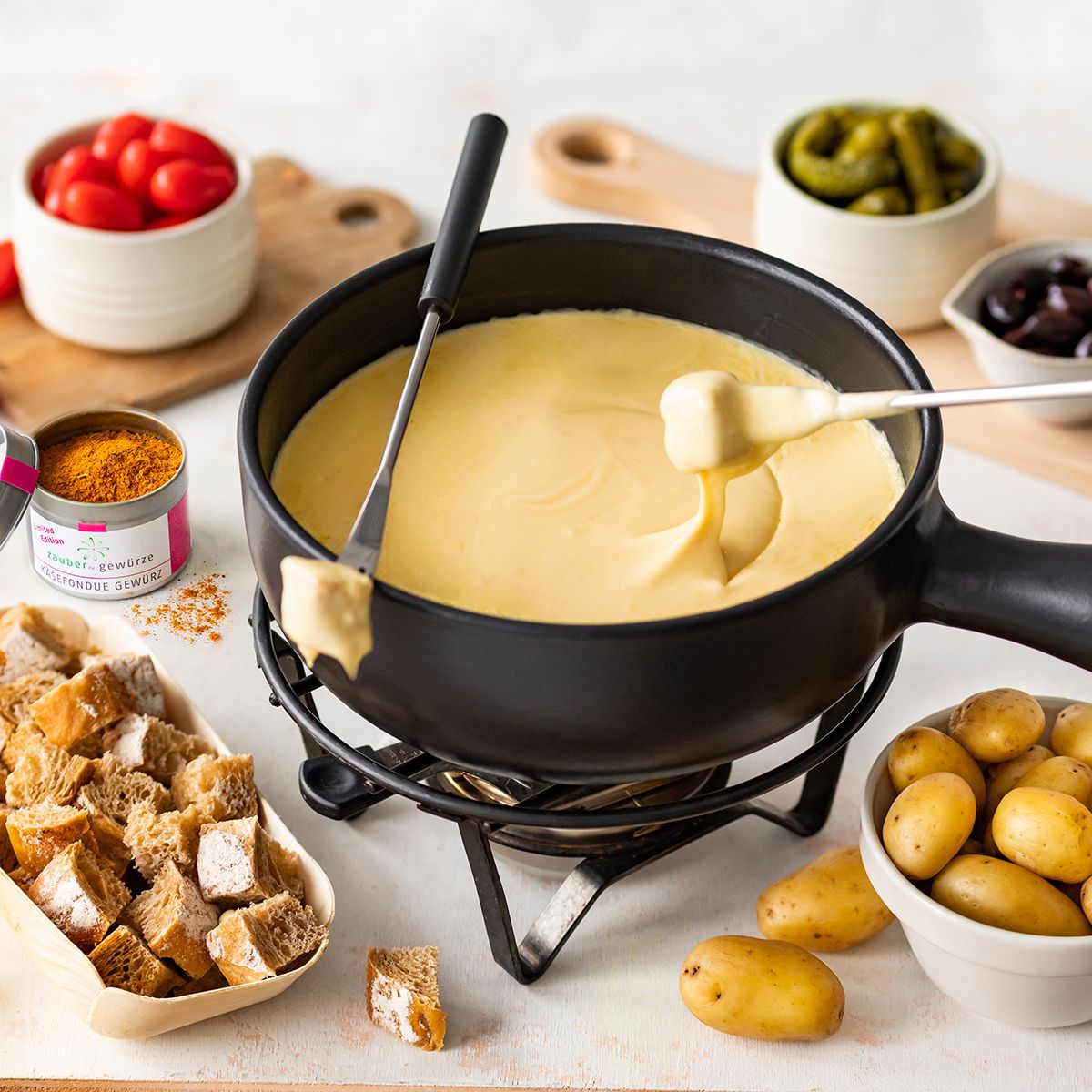 Käsefondue in schwarzem Topf. Brot, Kartoffeln, Gemüse und Gewürz daneben. Gabeln mit Käse.