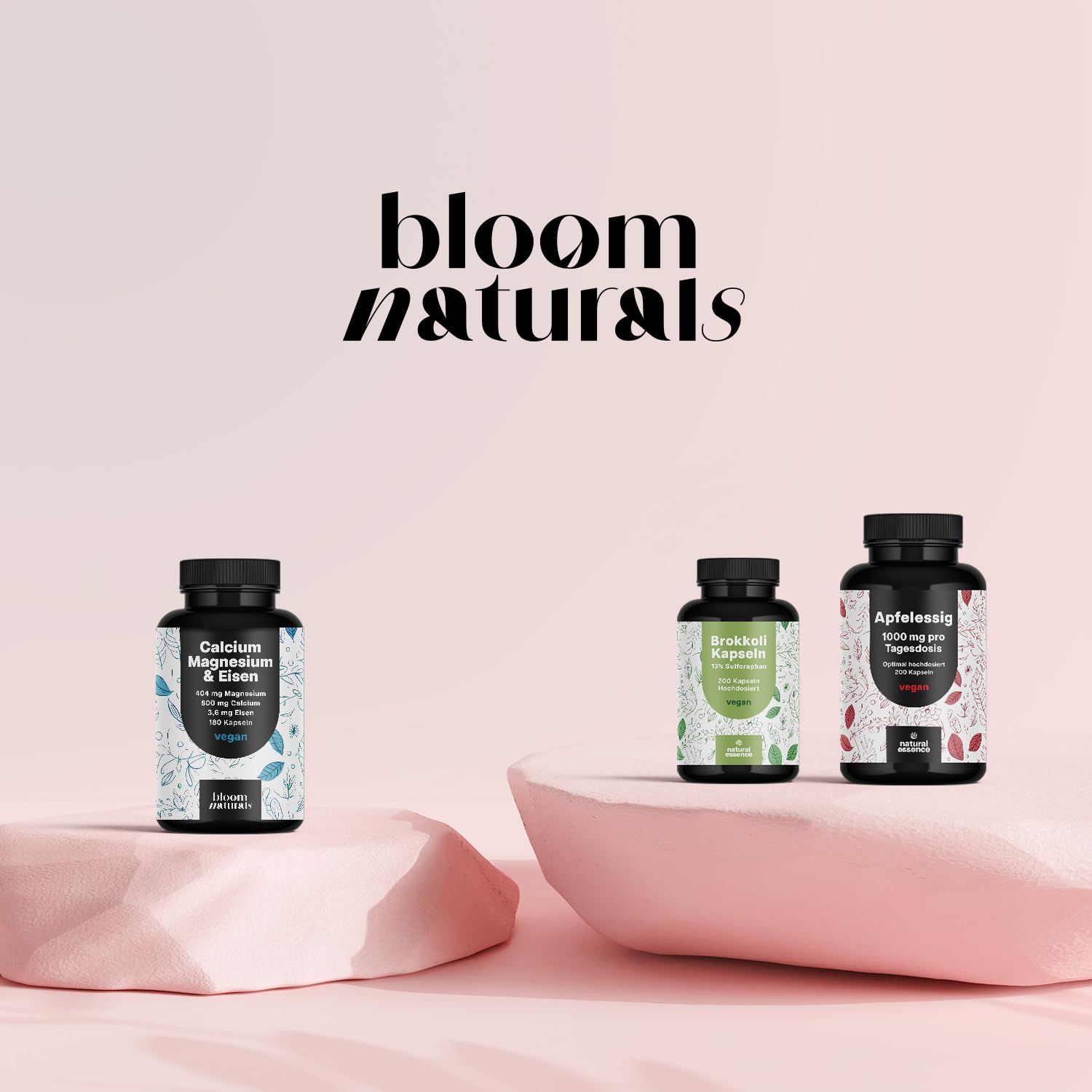 Drei Flaschen auf einer rosa Plattform. Marke: Bloom Naturals. Produkte: Calcium, Grüntee, Apfelessig.