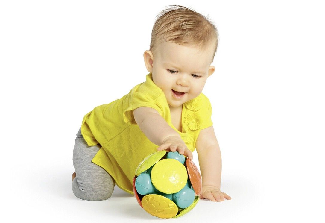 Baby krabbelt nach einem bunten Ball. Der Ball hat gelbe, blaue, orange und grüne Elemente. Das Baby trägt gelbes Shirt.