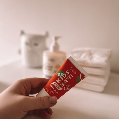 Hand hält Tube. Aufschrift: KIDS TOOTHPASTE, PREBIOTIC, NO FLUORIDE. Mit Illustrationen. Im Hintergrund Badutensilien.