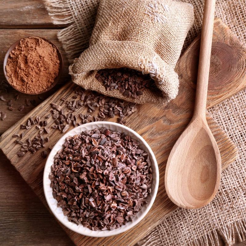 Draufsicht: Schüssel mit Kakaonibs, Schüssel mit Kakaopulver, Leinensack mit Nibs, Holzlöffel, Holzbrett, auf Holzuntergrund.