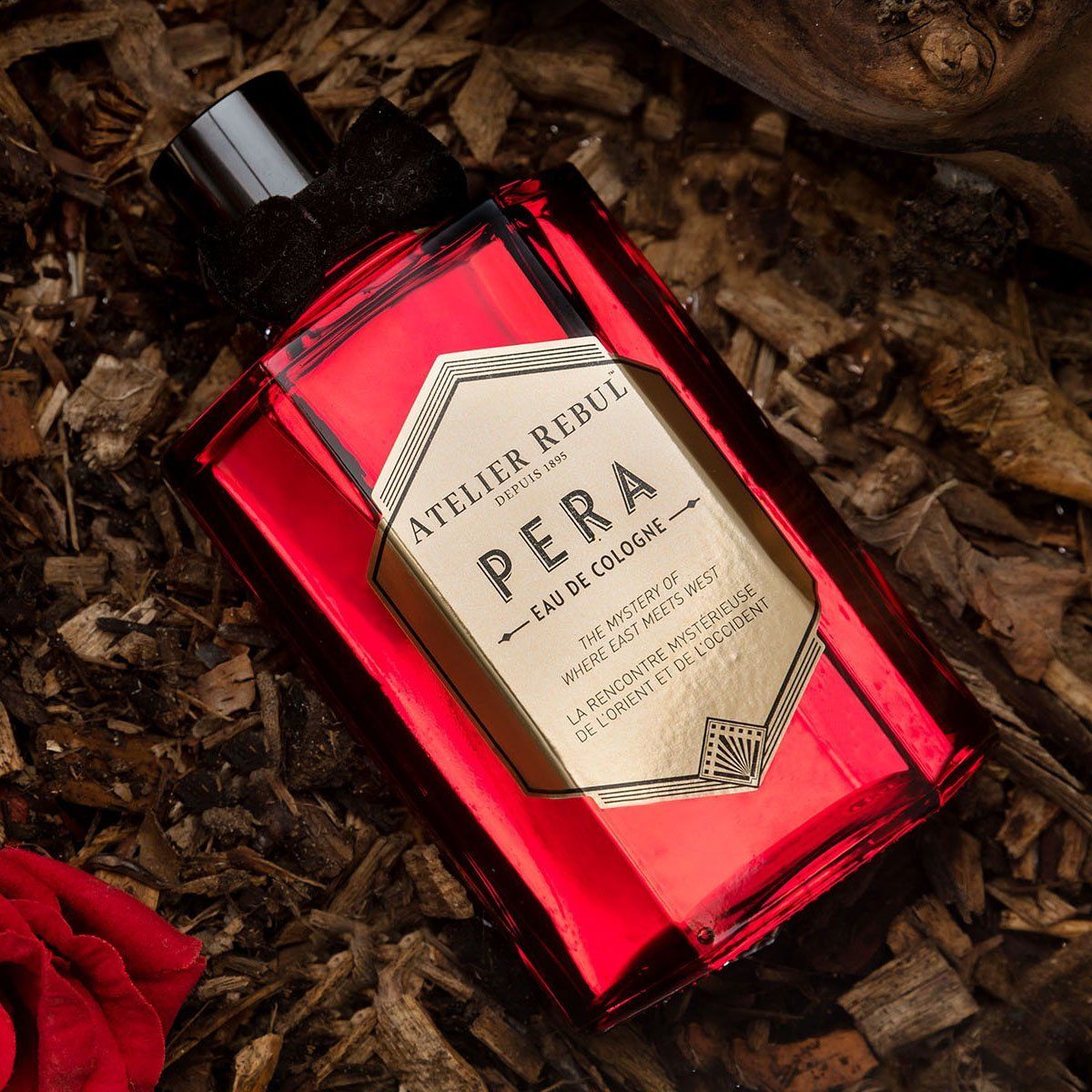 Rote Glasflasche mit schwarzem Verschluss und Schleife. Aufschrift: Atelier Rebul Pera Eau de Cologne. Auf Holzspänen platziert.