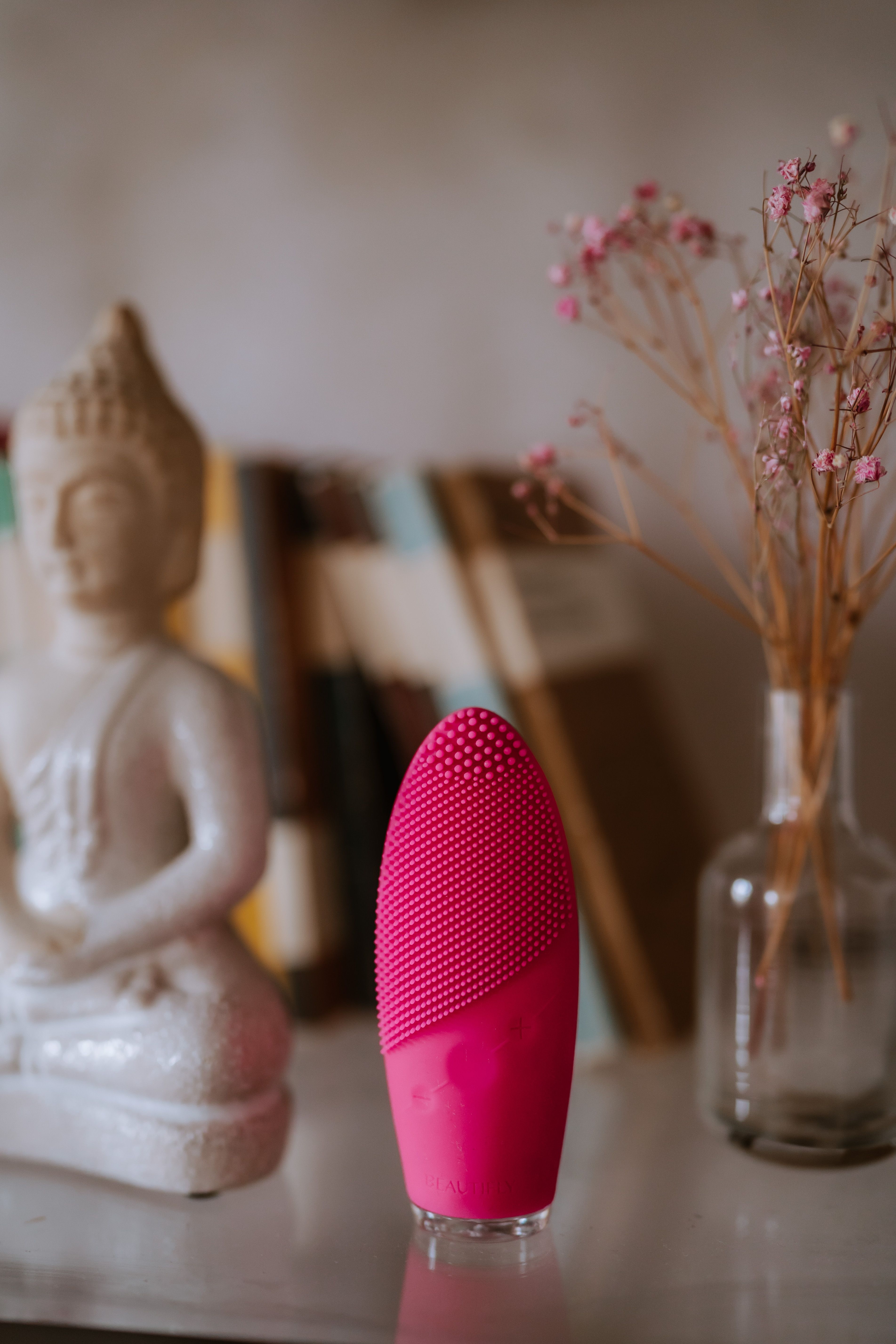 Pinkfarbene Schall-Gesichtsbürste steht auf einem Tisch. Buddha-Statue und Bücher im Hintergrund.