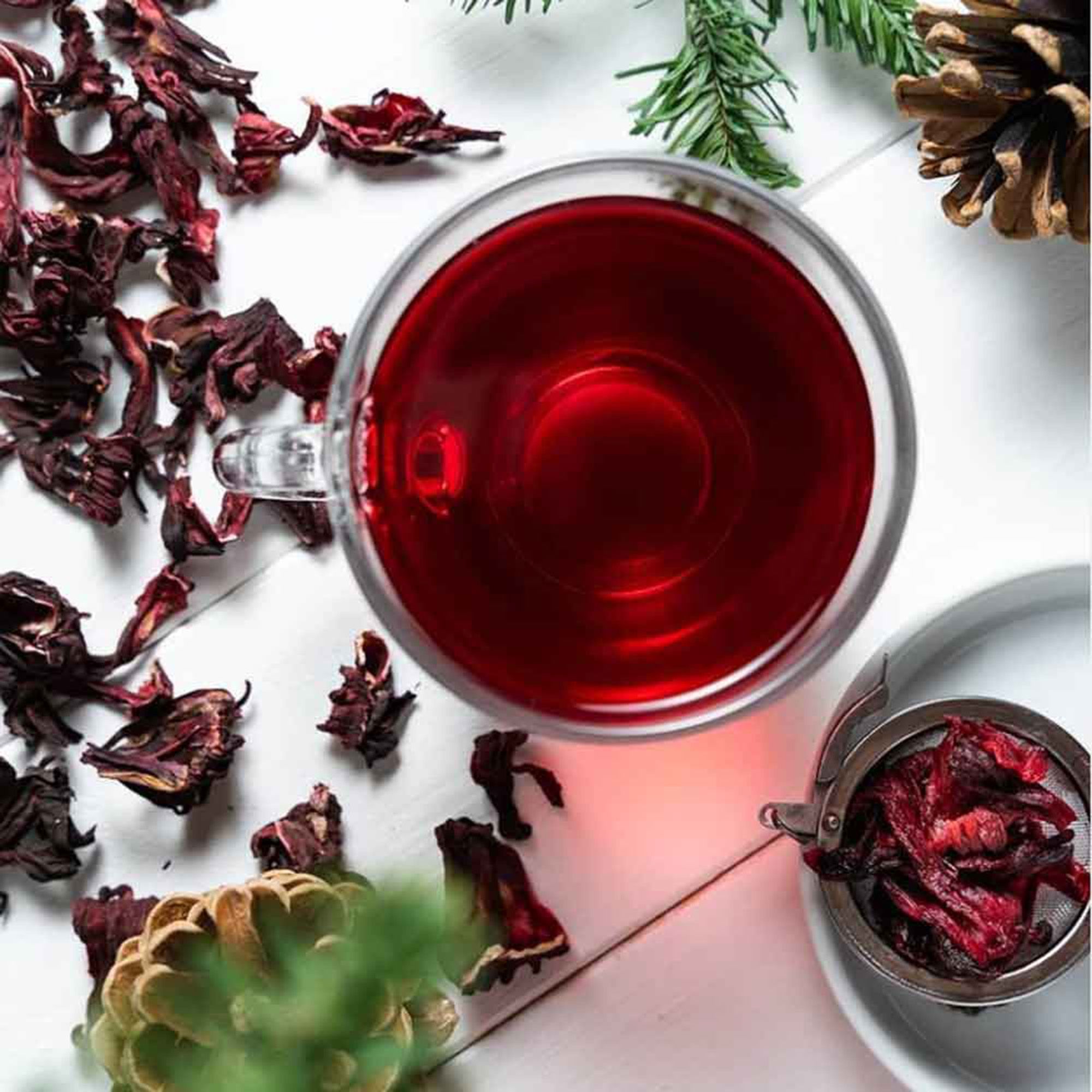 Eine Tasse mit rotem Hibiskustee, daneben getrocknete Blüten und ein Teesieb.