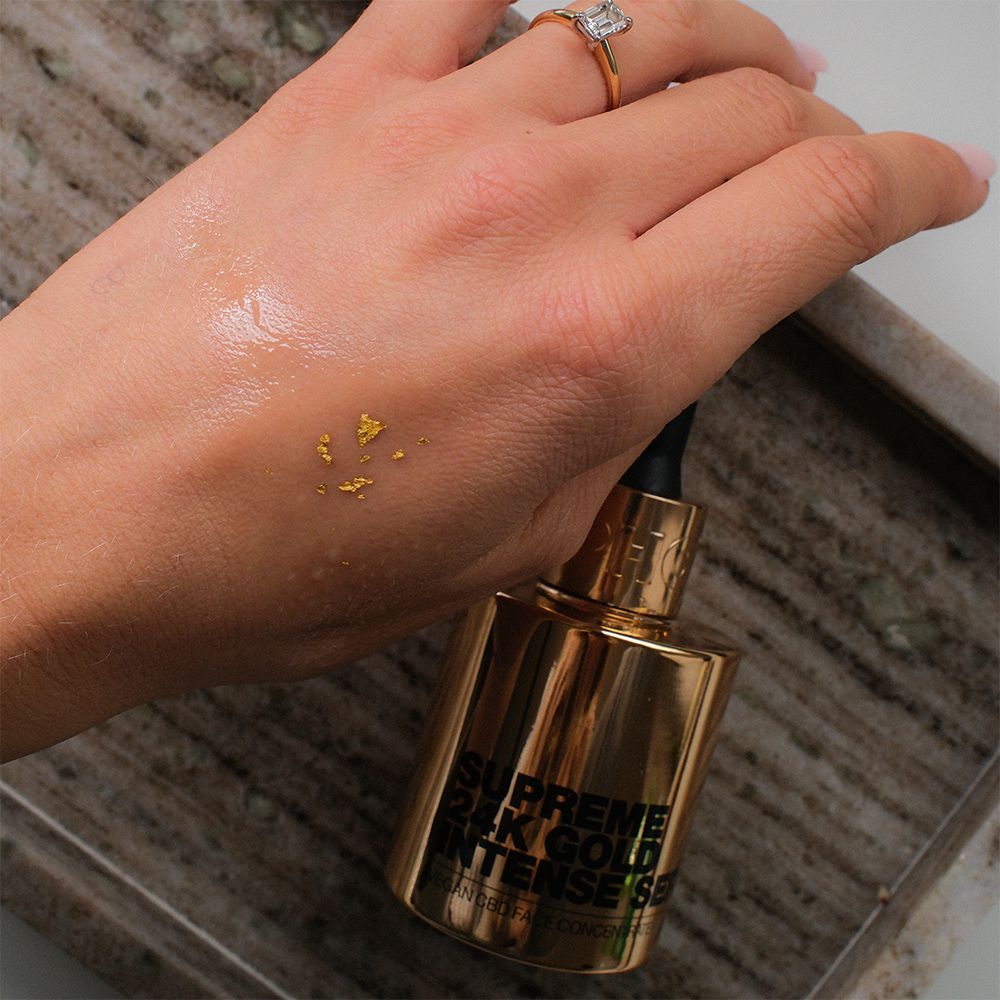 Hand mit Serum und Goldflocken. Goldene Serumflasche mit Pipette. Aufschrift: Supreme 24K Gold Intense Serum.