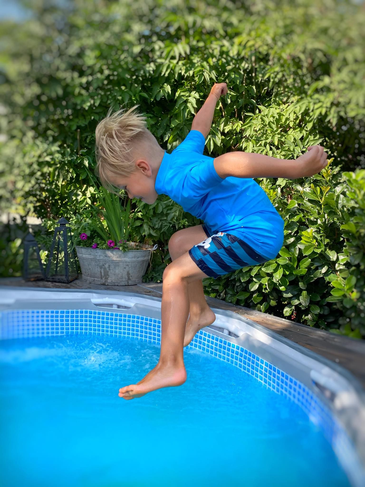 Kind springt in einen Pool, trägt blaues Shirt und Badehose. iQ-UV-Logo auf dem Shirt. Grüne Umgebung.