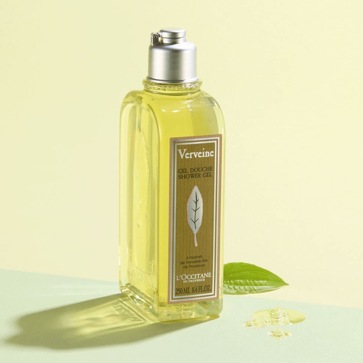 Duschgel-Flasche mit silbernem Deckel. Aufschrift "Verveine". Blatt-Dekor. L'Occitane-Logo. Gelber Hintergrund.