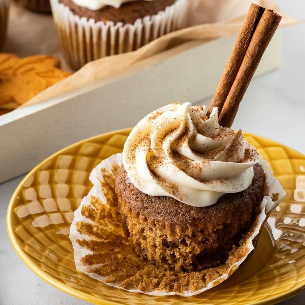 Muffin mit Sahnehaube und Zimtstangen auf gelbem Teller. Papierförmchen. Im Hintergrund weitere Backwaren.