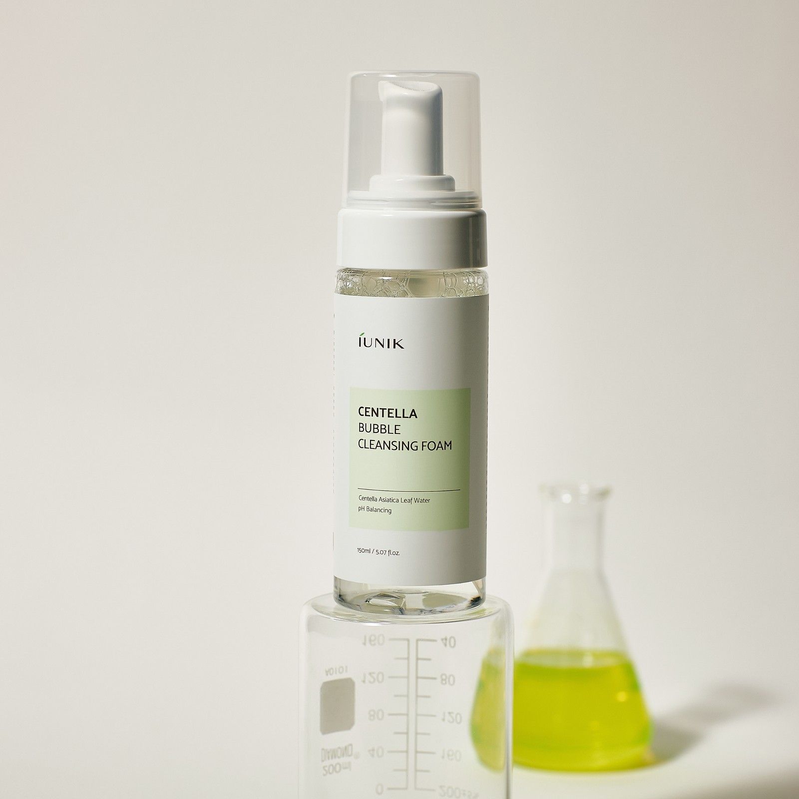 iUNIK Centella Bubble Cleansing Foam Flasche auf einem Messzylinder. Ein Becherglas mit gelber Flüssigkeit steht daneben.